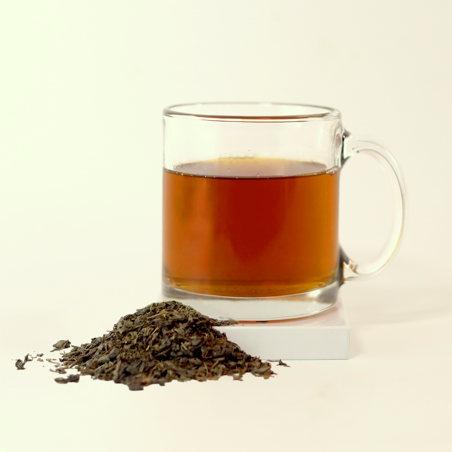  A clear glass mug filled with orange-brown tea - Moonlit Mint -  next to a small pile of dried tea leaves. A Good Store product.