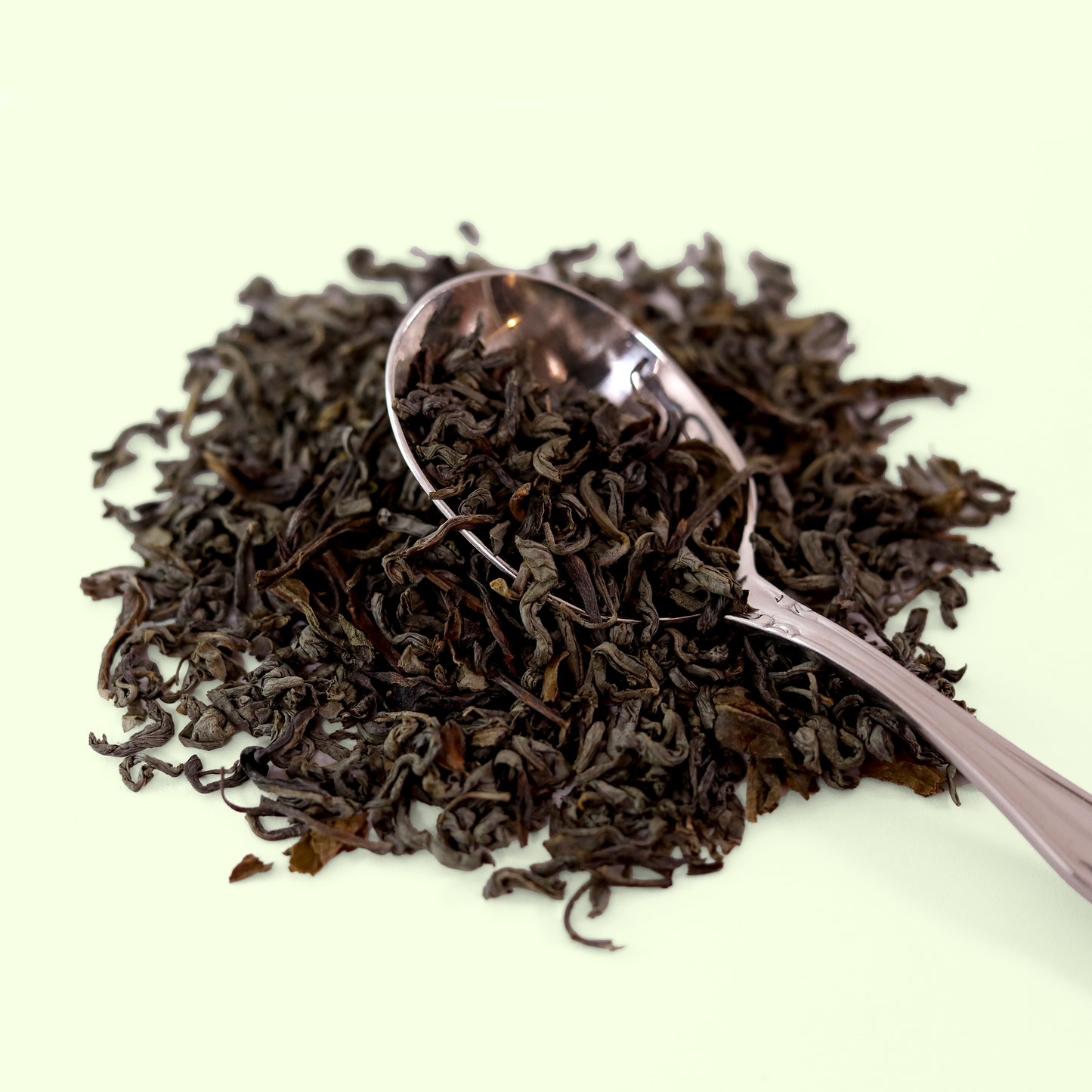  A close-up view of a pile of dried April Meadows Green tea leaves. A metal spoon is visible, scooping up some of the tea blend. A Good Store product.