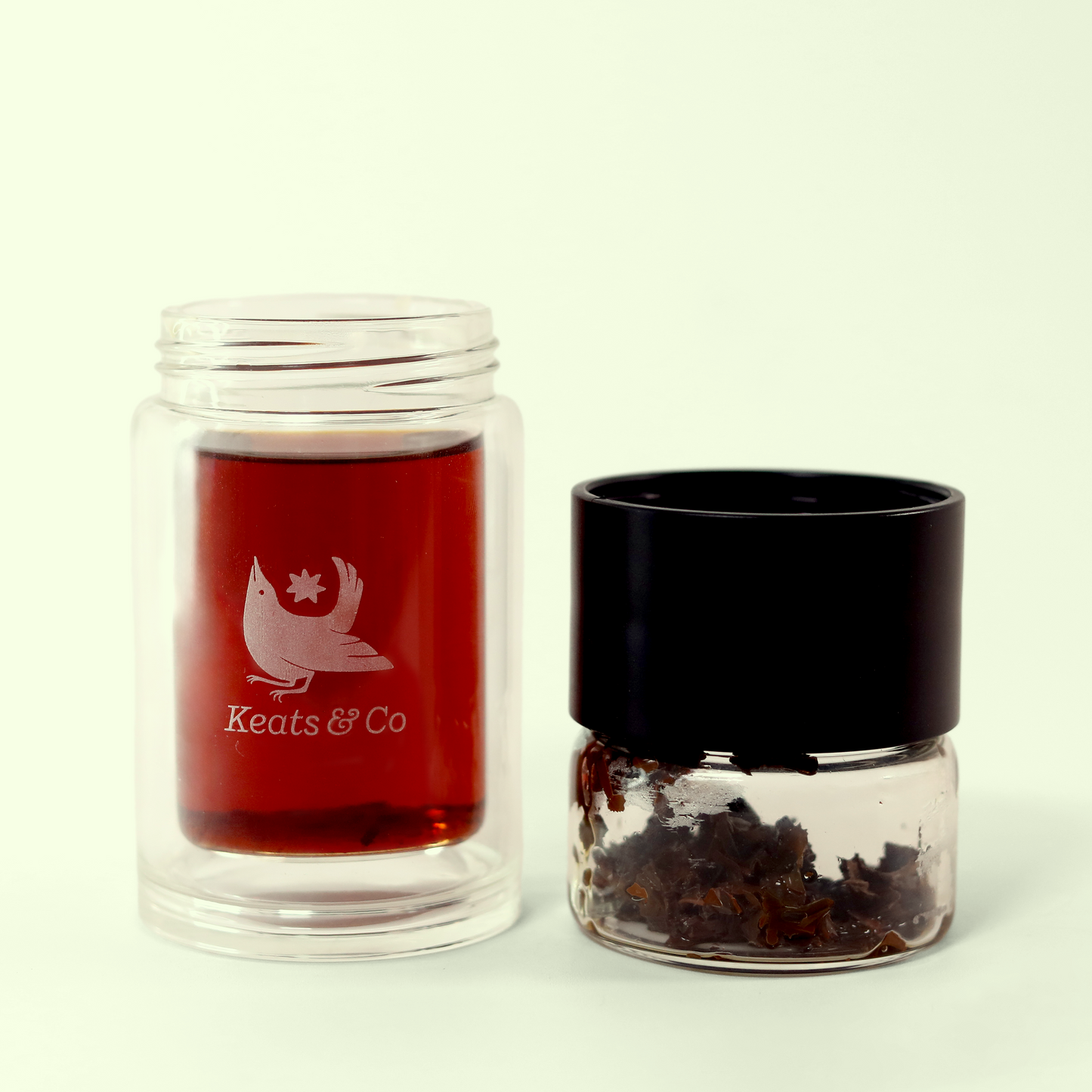 A clear glass double-walled jar with tea inside, sits next to a small clear glass jar filled with dark, wet tea leaves. The glass jar with the tea has a white label featuring a nightingale and the text "Keats & Co" engraved on it. A Good Store product.