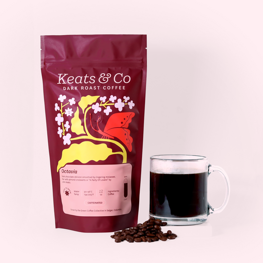 A burgundy-colored coffee bag labeled "Keats & Co Dark Roast Coffee" with colorful floral and butterfly designs. Next to the bag is a clear glass mug filled with dark coffee and a few coffee beans scattered around. The items are placed on a white surface against a light pink background. A Good Store product.