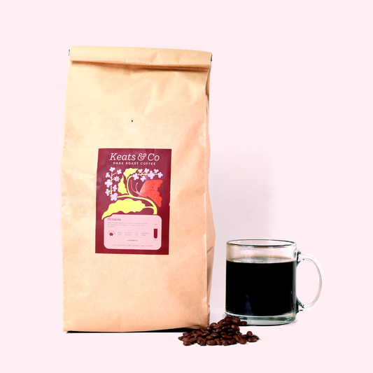 A large tan paper bag of Keats & Co Dark Roast Coffee with a burgundy label featuring floral and butterfly designs. Next to the bag is a clear glass mug filled with black coffee, and a small pile of whole coffee beans in front. The items are arranged on a white surface against a light background. A Good Store product.