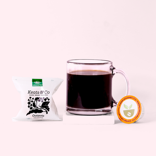 A glass mug filled with dark coffee, flanked by a white Keats & Co coffee packet on the left and a single-serve coffee pod with an orange label on the right, all arranged on a white surface against a light pink background.