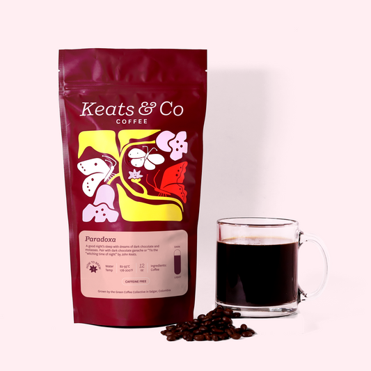 A burgundy-colored coffee bag labeled "Keats & Co Dark Roast Coffee-Paradoxa" with colorful floral and butterfly designs. Next to the bag is a clear glass mug filled with dark coffee and a few coffee beans scattered around. The items are placed on a white surface against a light pink background. A Good Store product.