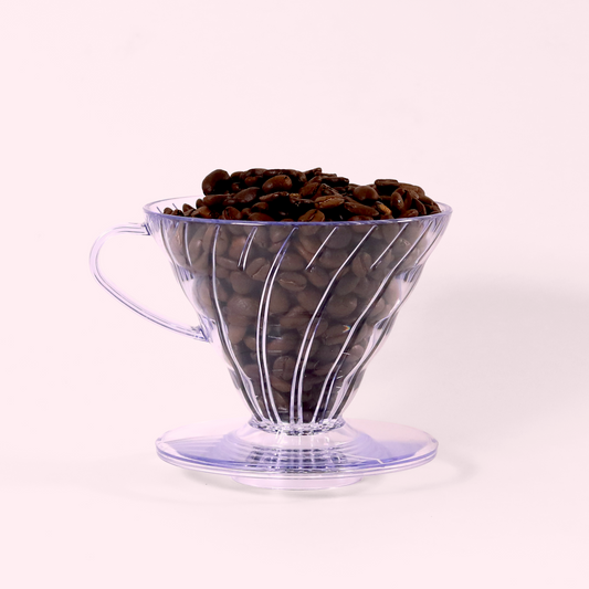 A close-up view of a clear plastic pour-over coffee dripper filled with dark roasted coffee beans. The dripper has a conical shape with spiral ridges and sits on a circular base.