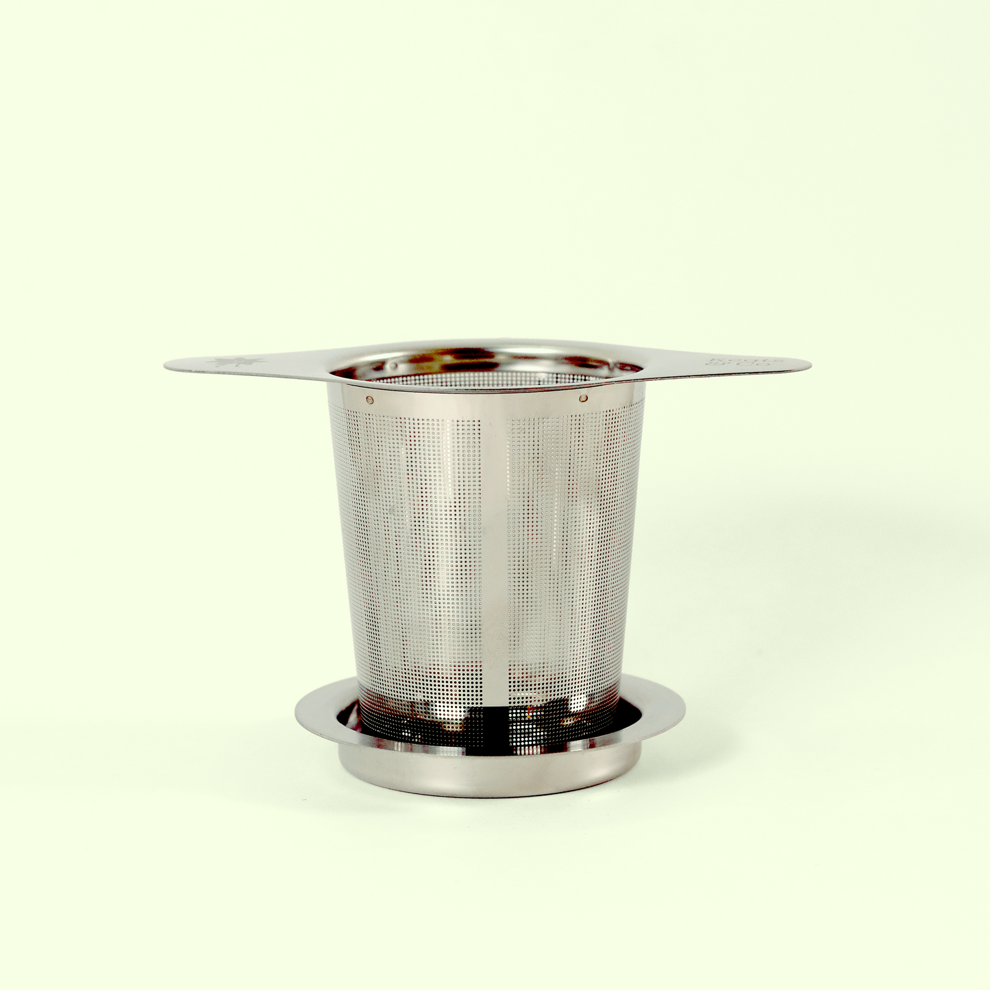 A stainless steel tea infuser with a perforated mesh basket and wide rim, sitting on a small base. The infuser is designed to hold loose tea leaves while brewing.