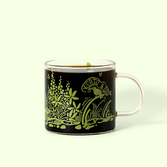 A detailed view of the Keats & Co Meadow glass mug. It's filled with coffee and the mug's design shows green illustrations of plants, flowers, and a bird, along with the Keats & Co logo. A Good Store product. 