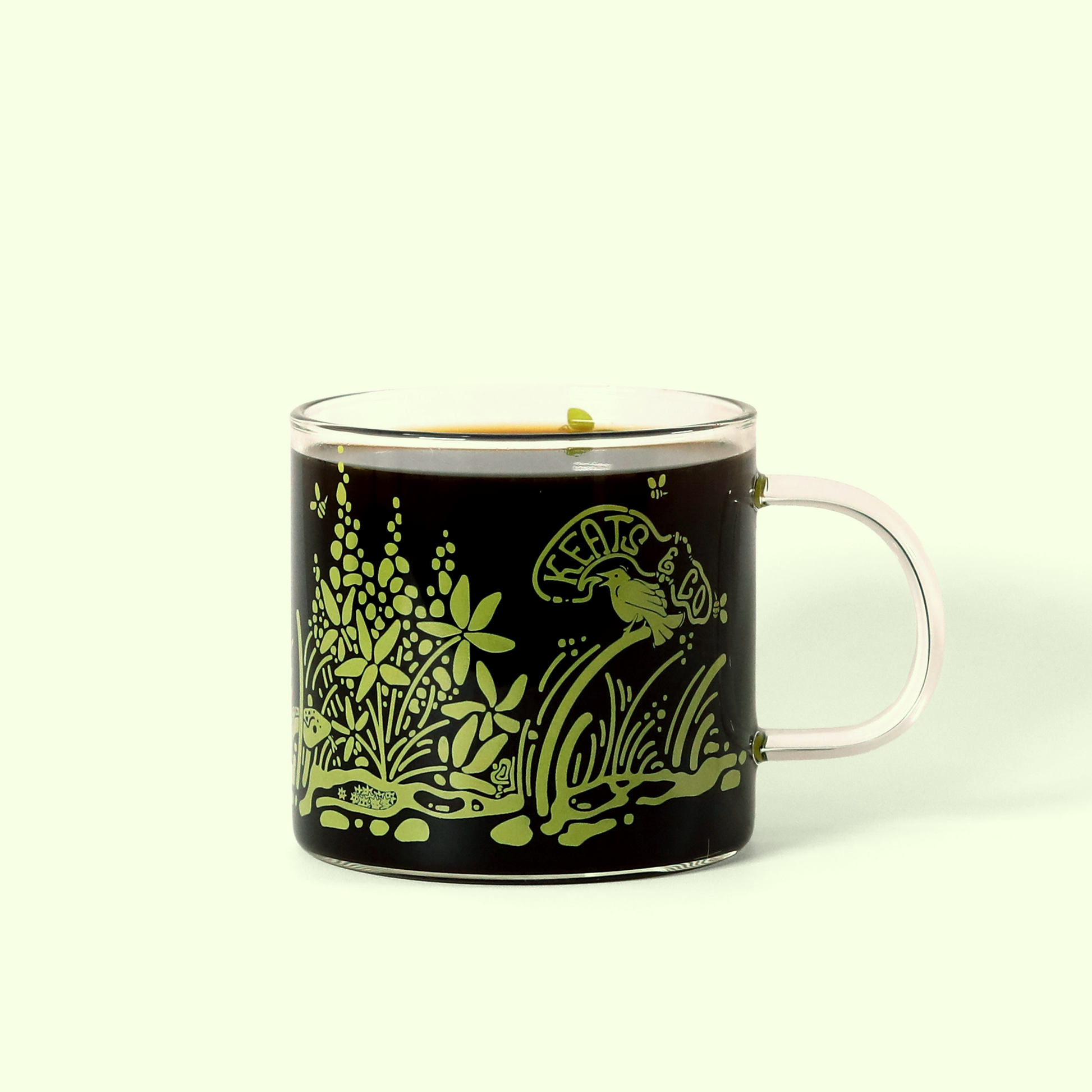 A clear glass coffee mug with a black liquid inside, decorated with lime green nature-inspired designs including leaves, flowers, and a bird. The decorative pattern creates a garden scene around the mug's exterior.