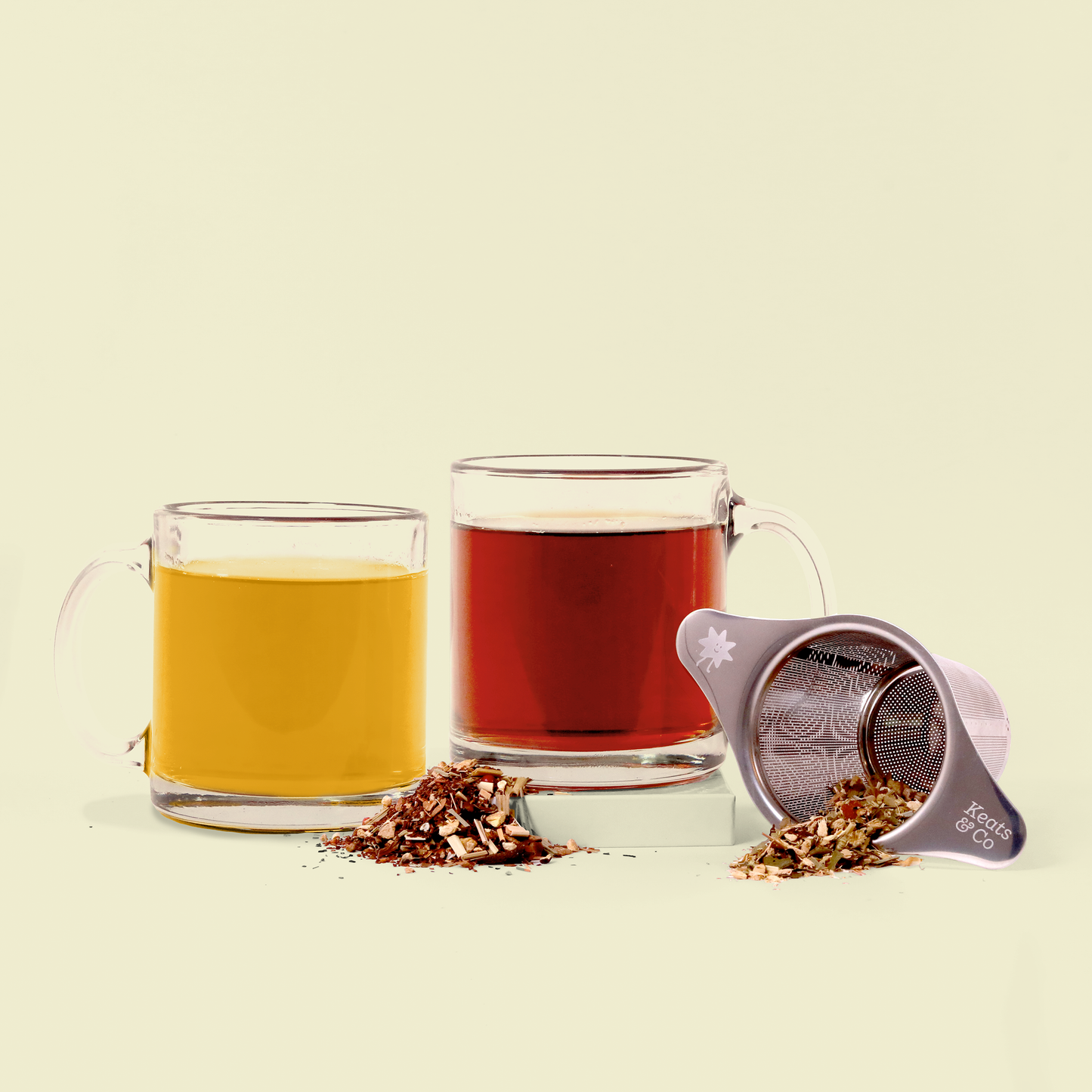 Two clear glass mugs with different teas - one golden yellow and one deep red - displayed alongside loose herbal tea blend and a silver Keats & Co tea strainer against a cream background. 