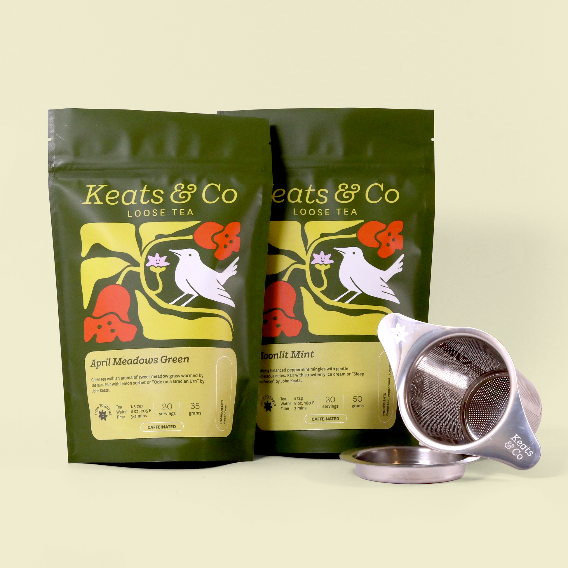 Product photo of two Keats & Co loose tea packages in dark green featuring a white bird and floral design. The varieties shown are "April Meadows Green" and "Moonlit Mint." A silver Keats & Co tea strainer with its base is displayed alongside the packages against a cream background.