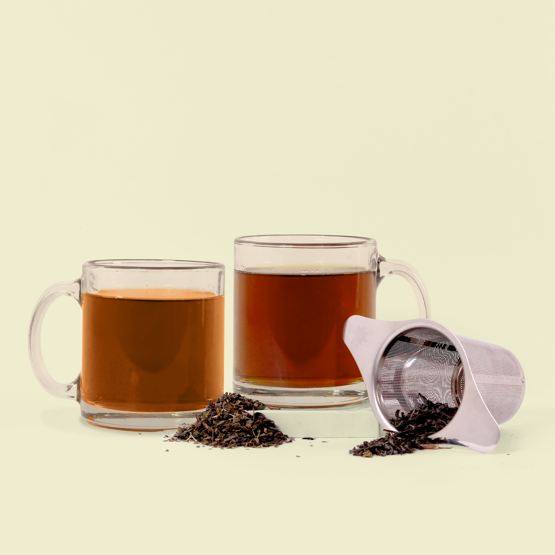 Two glass mugs filled with different colored teas (one lighter and one darker amber) displayed with loose tea leaves scattered in front and a silver tea strainer tipped over, spilling some tea leaves. The scene is photographed against a cream-colored background.