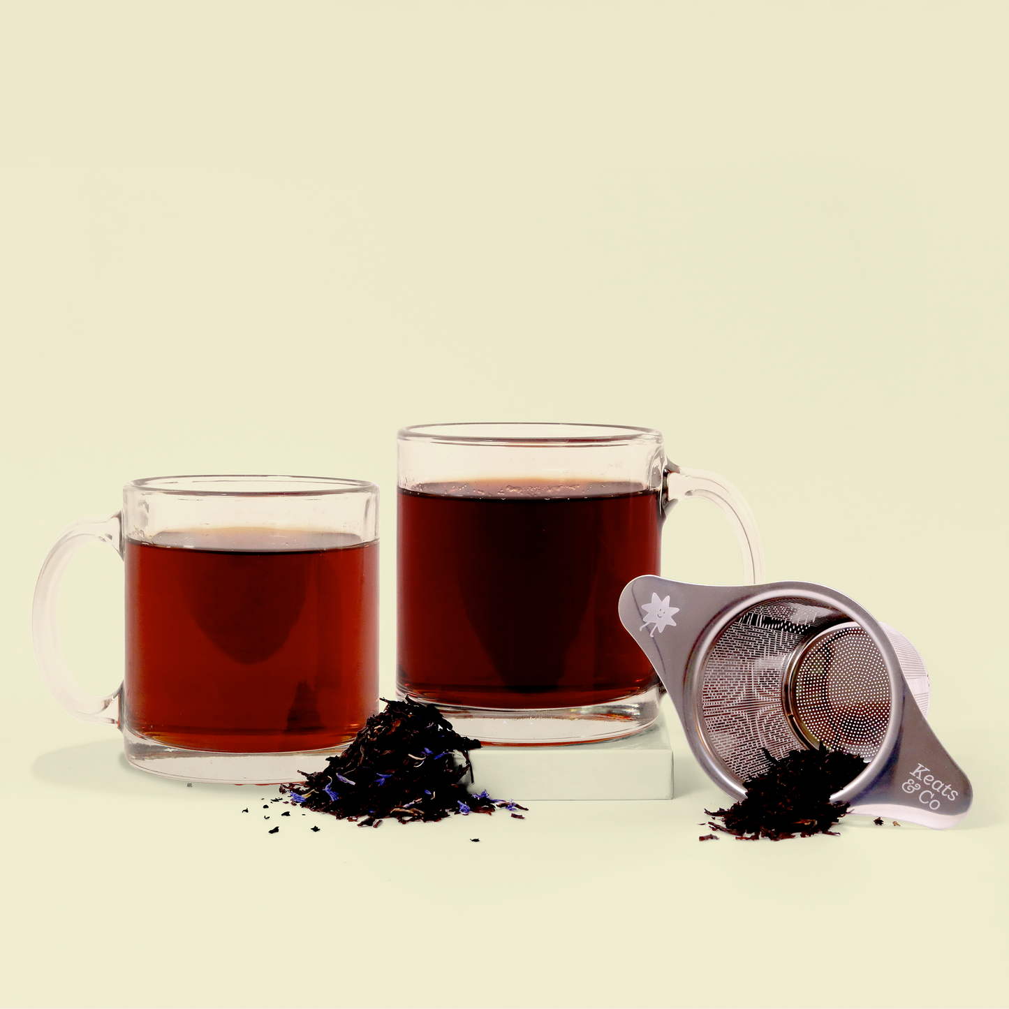 Two clear glass mugs filled with dark red tea, alongside loose tea leaves and a silver Keats & Co tea strainer against a cream-colored background. 