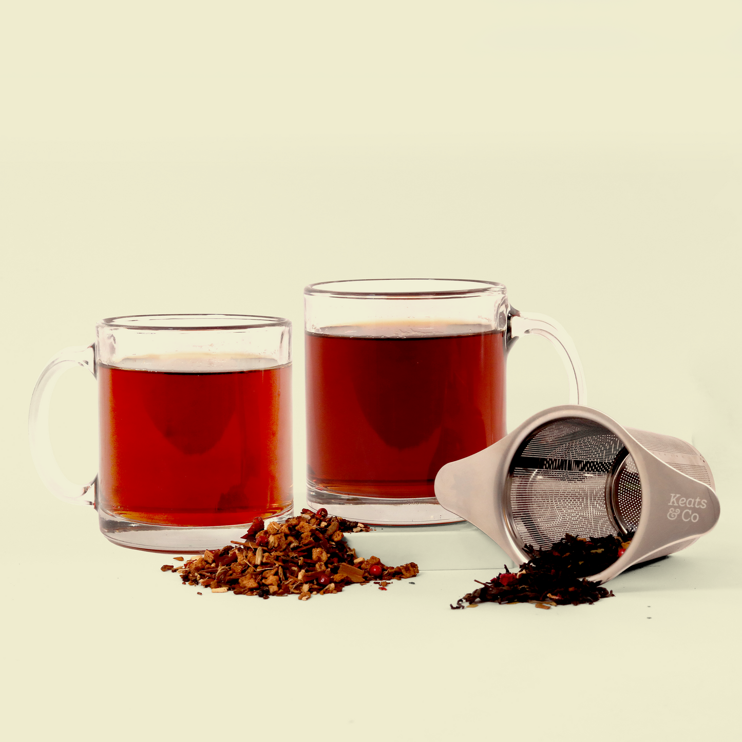 Two clear glass mugs filled with reddish-brown tea, alongside loose tea leaves scattered on a white surface. A metal Keats & Co tea strainer is tipped over, spilling some loose tea leaves.