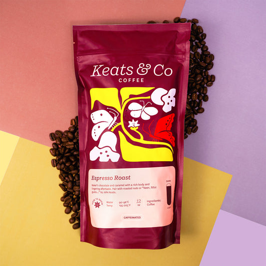 A stand-up pouch of Keats & Co Coffee Espresso Roast, styled on a geometric pastel background with scattered coffee beans. The pouch is deep burgundy with a yellow, white, and red graphic design and a light pink label at the bottom.