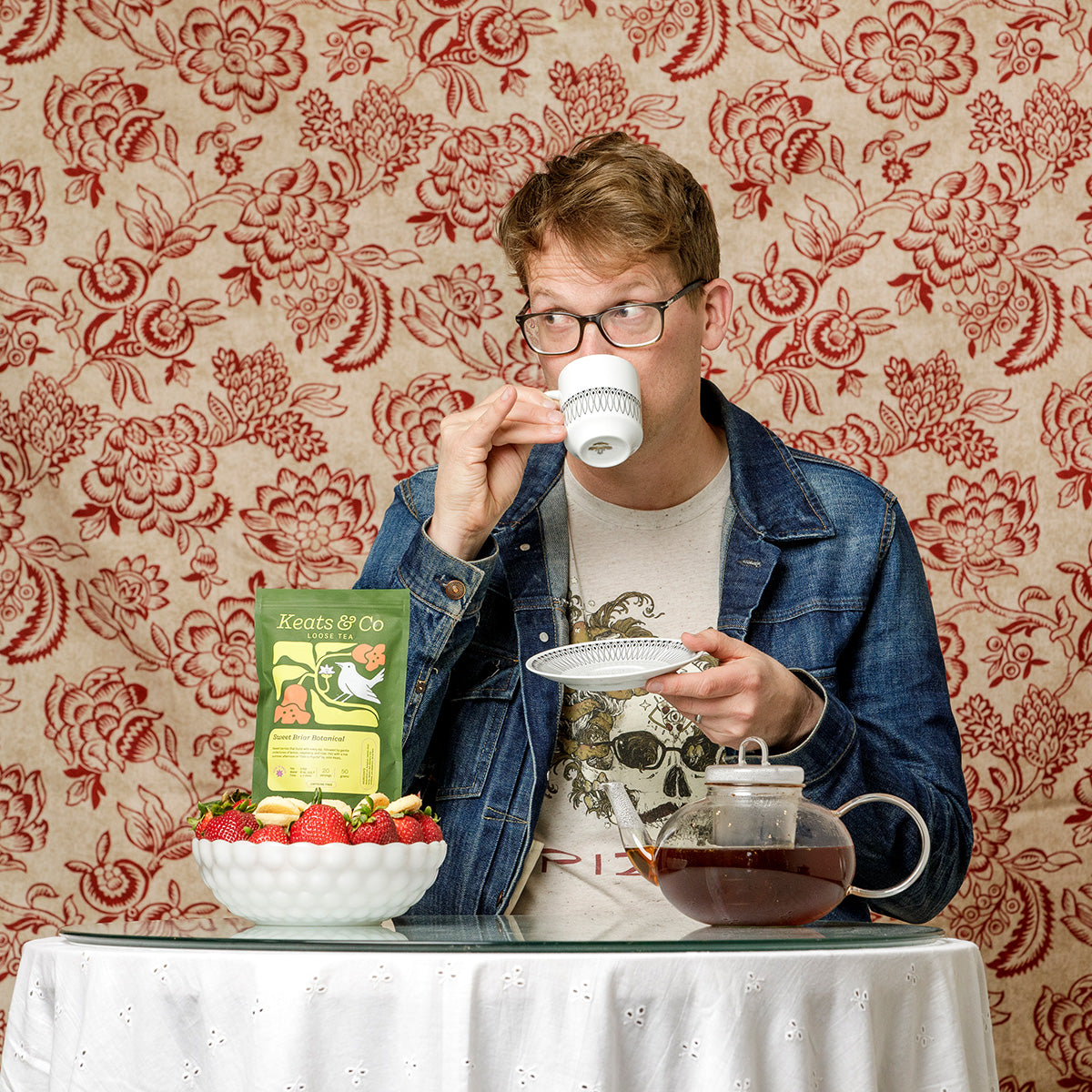 Hank Green sitting at a white lace-covered table, drinking tea from a delicate white teacup and saucer. On the table are fresh strawberries in a white bowl, a glass teapot with dark tea, and a green package of "Keats & Co" tea. The background features an ornate red and cream floral wallpaper pattern. The scene has a cozy, vintage tea time aesthetic.