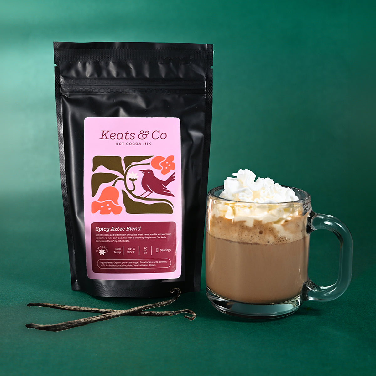 A black stand-up pouch of Keats & Co Spicy Aztec Blend hot cocoa mix stands next to a clear glass mug filled with a hot cocoa drink topped with whipped cream, on a dark green background with vanilla beans in front.