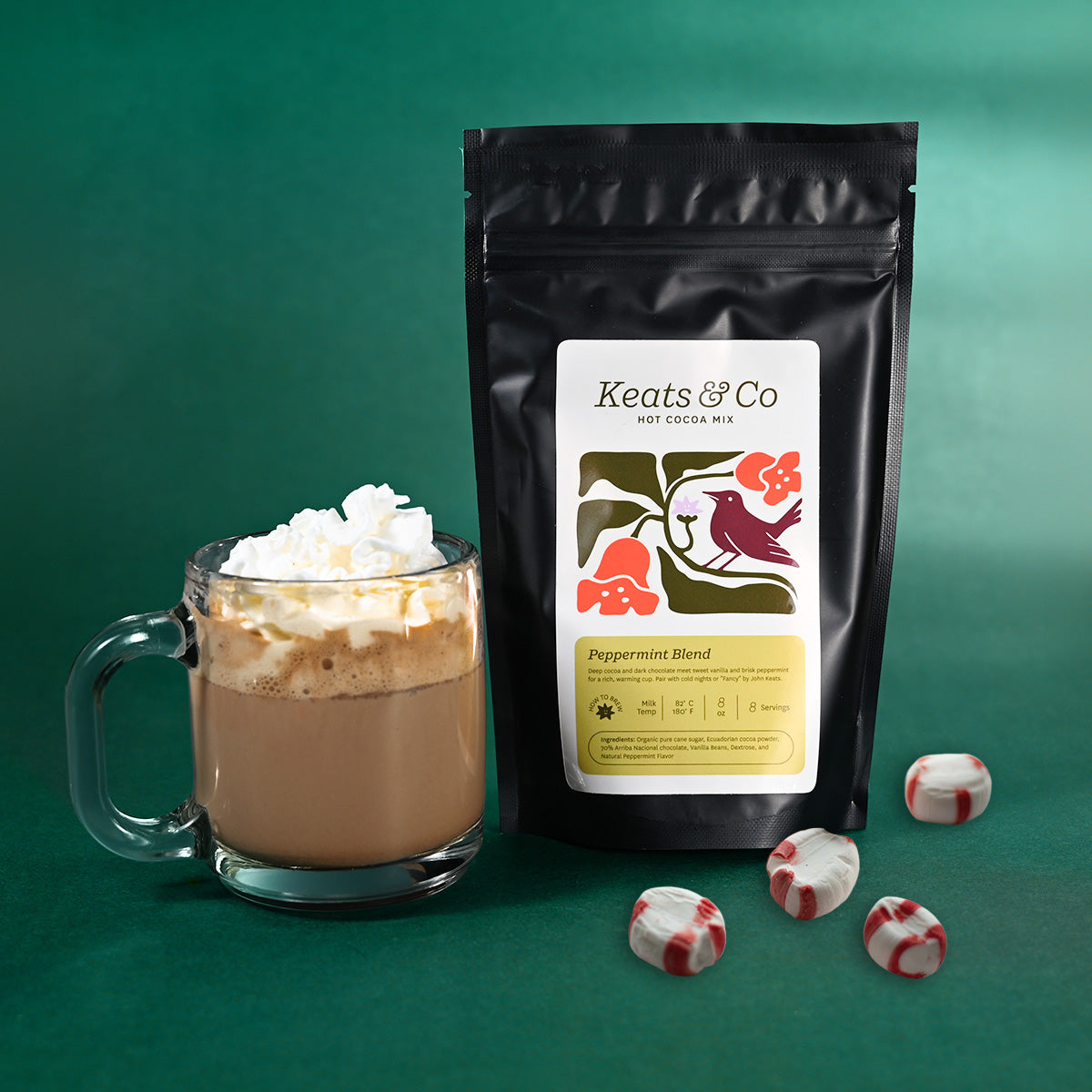 A black stand-up pouch of Keats & Co peppermint blend hot cocoa mix stands next to a glass mug filled with hot cocoa topped with whipped cream, with a few peppermint candies on a green background.