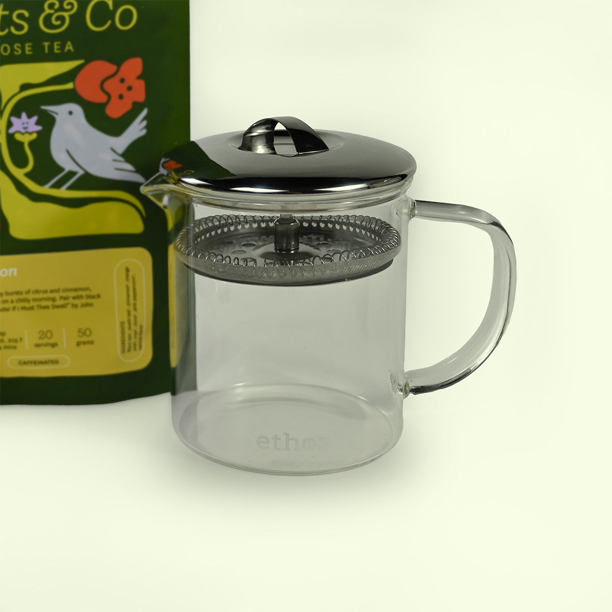 Clear glass tea press with metal lid and handle, next to a box of tea on a light background