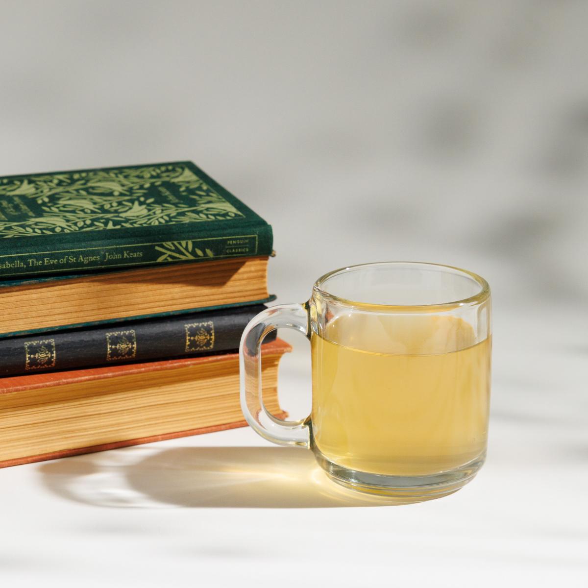 Cup of yellowish Keats & Co Loose Leaf Thrushsong Oolong tea in a glass cup next to a pile of John Keats books on a white background. 