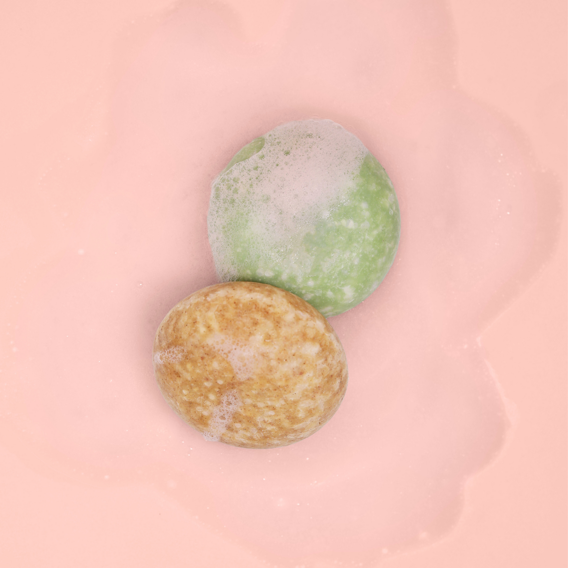 The Dew Drop and Bloom Shampoo bars covered in soap on a pink background. 
