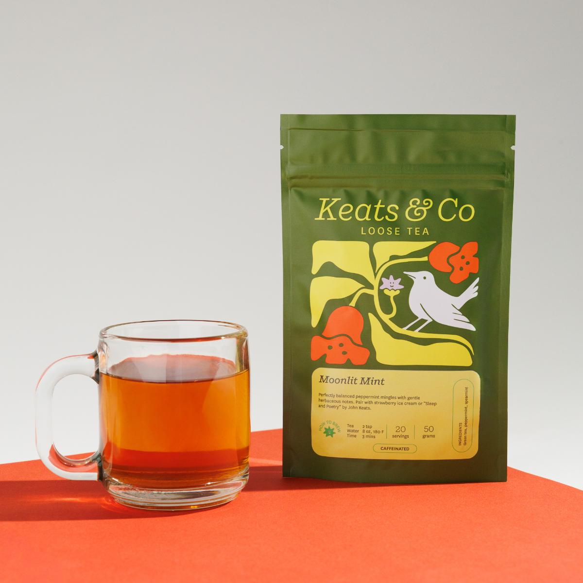 Green pouch of Keats & Co Loose Tea Moonlit Mint against a white background, featuring illustrated artwork of a white bird and orange flowers, displayed next to a clear glass mug filled with yellowish brewed tea on an orange surface.