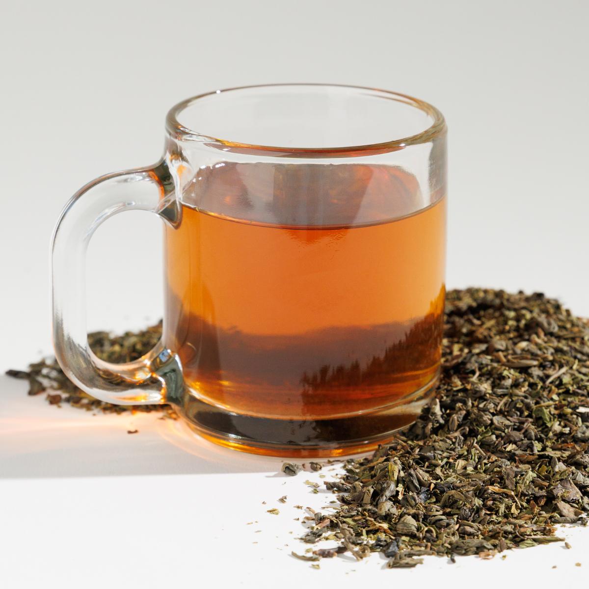 Glass cup of yellowish Moonlit Mint tea positioned next to a pile of loose leaf tea blend on a reflective white surface against a soft white background. A Good Store product.