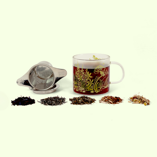 A glass mug with green botanical illustrations containing brewed tea, a metal tea infuser with saucer, and five small piles of different loose tea varieties arranged in a row, showing the progression from dark to light tea blends.
