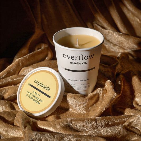 A white candle jar labeled 'overflow candle co.' with its lid placed beside it on draped metallic gold fabric.