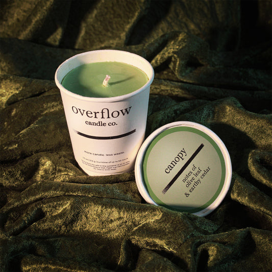 A white candle jar labeled 'overflow candle co.' with pale green wax sits beside its matching lid, which reads 'canopy' and 'moss, sage, & earthy scents,' set on draped dark olive fabric.