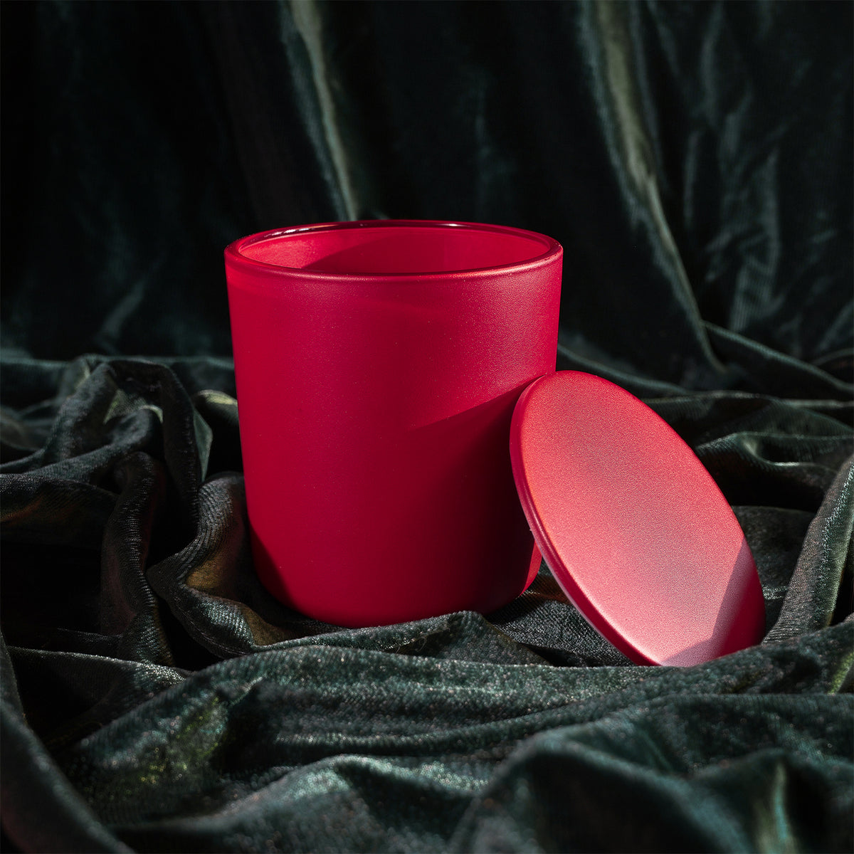 A bright red cylindrical candle jar with its matching lid resting beside it, set against a draped black velvet fabric background.