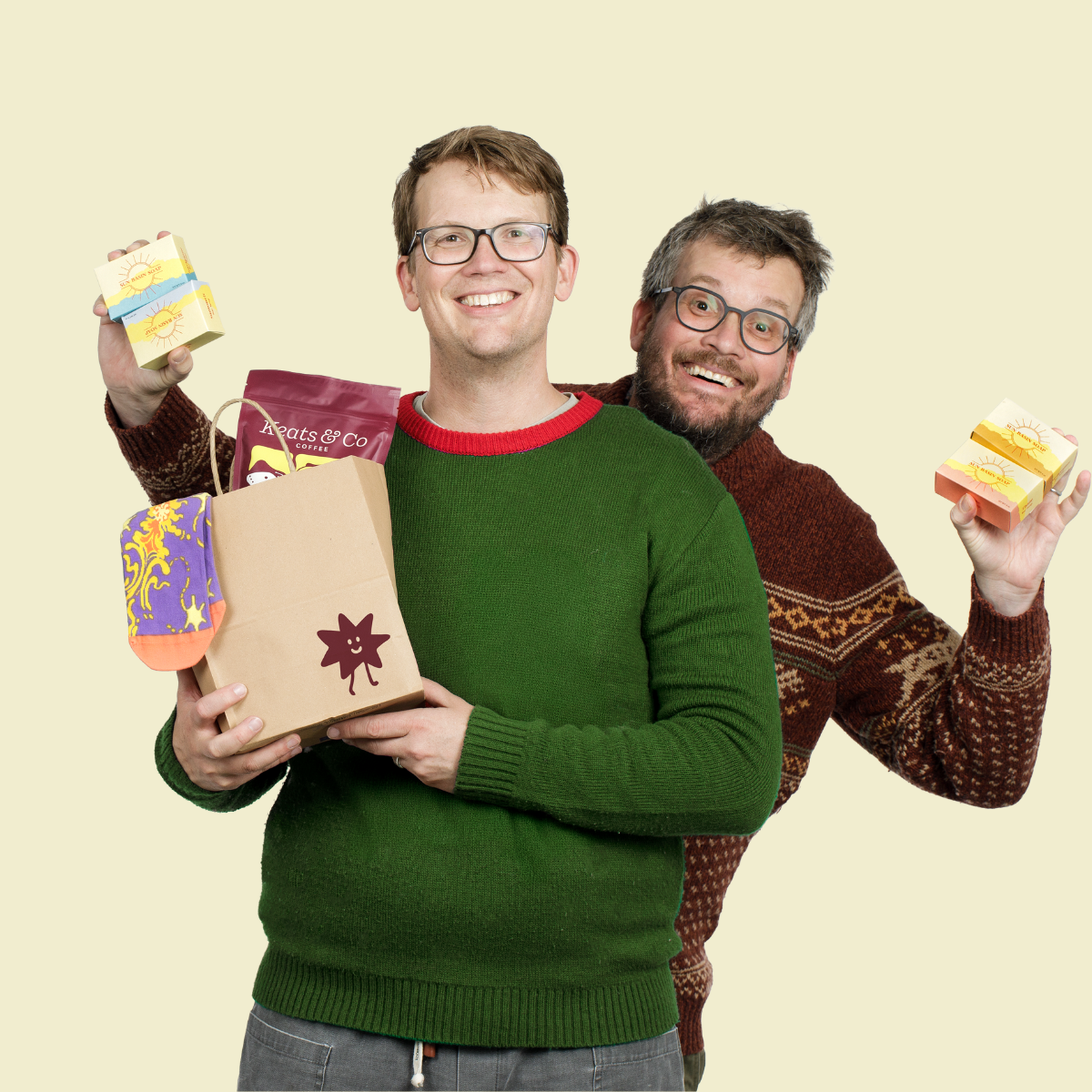Hank and John Green holding various products and a bag with the Good Store logo on it against a beige background