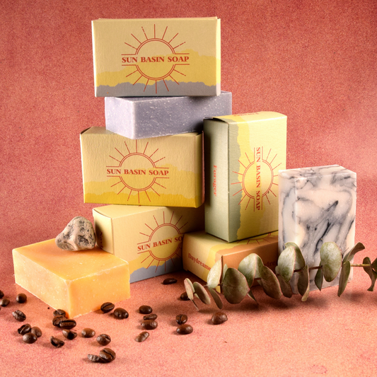A photo of soaps stacked on top of each other, some in boxes with the Sun Basin Soap logo on them and some showing the color of the soap. The stack of soap is in front of a terracotta background with coffee beans and a eucalyptus surrounding it. A Good Store subscription product.