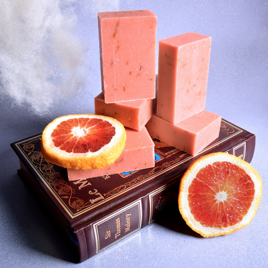 Five pink bars of soap are stacked on top of a hardcover book with two slices of Blood Orange and a puff of cloud behind it. The bars of soap are in front of a lavender background. The Daydreamer soap from Sun Basin Soap. A Good Store subscription product.