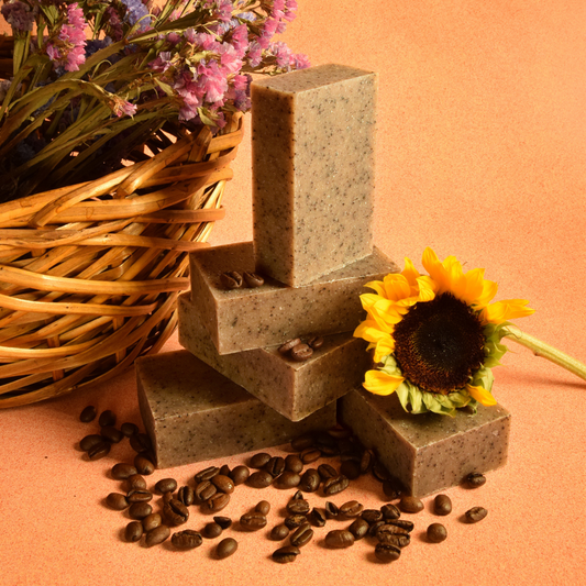 Five brown speckled bars of soap are stacked on top of each other surrounded by coffee beans, with a sunflower and a basket of flowers resting next to them. The bars of soap are in front of a light orange background. The Forager soap from Sun Basin Soap. A Good Store subscription.