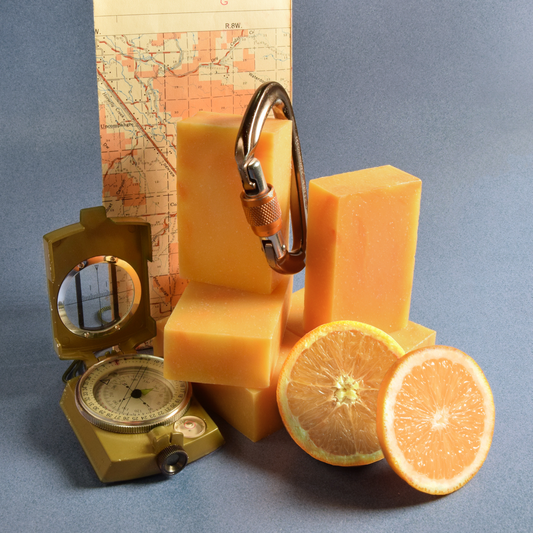 Five orange bars of soap are stacked on top of each other with a carabiner hanging from one of the soaps. A mar, compass and two grapefruit slices lay next to the soap. The bars of soap are in front of a light blue background. The Trailblazer soap from Sun Basin Soap. A Good Store subscription product.