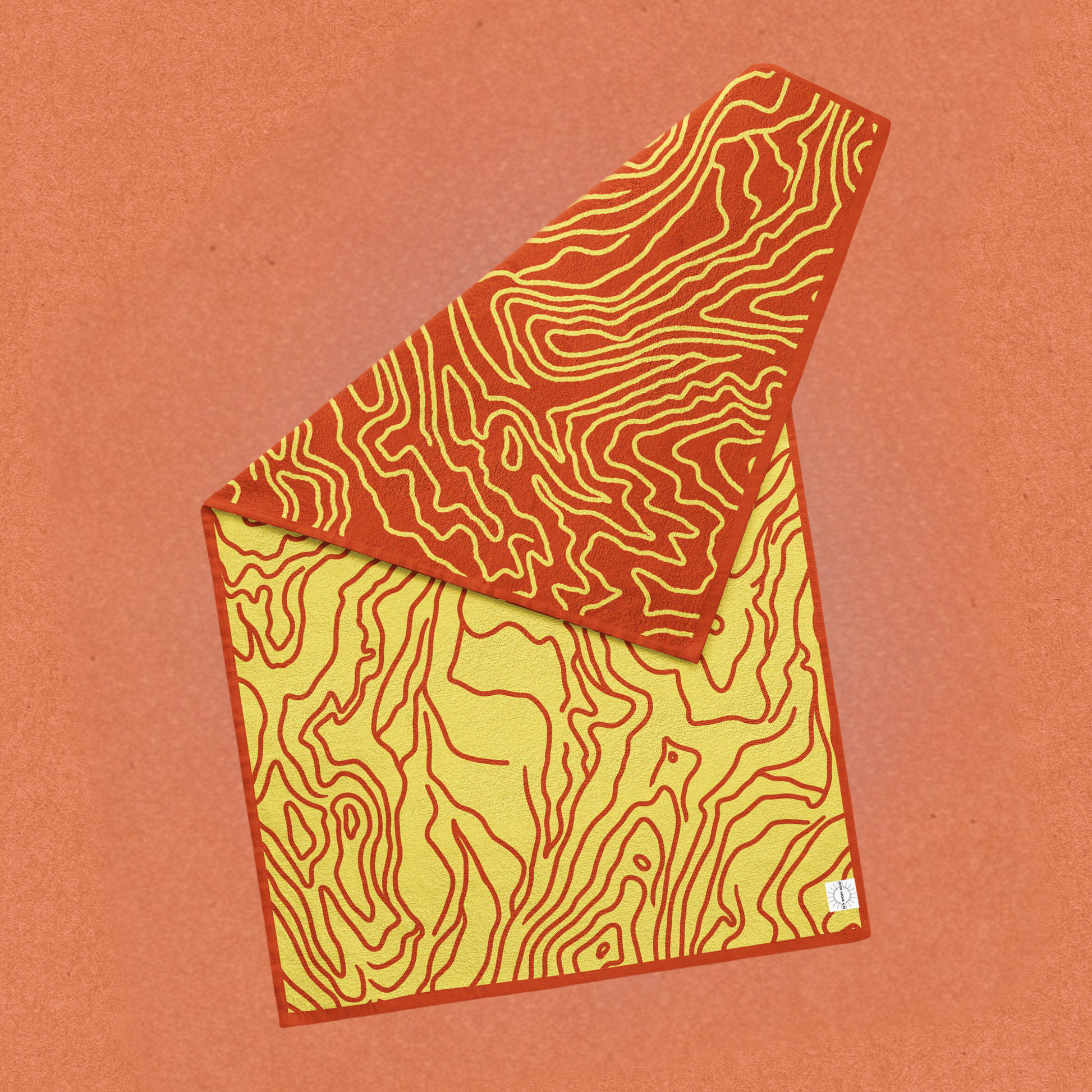 A reversible dark orange and yellow topographic towel with a corner folded down with a small tag that has the Sun Basin Soap logo. The towel is in front of a light orange background. A Good Store subscription product.