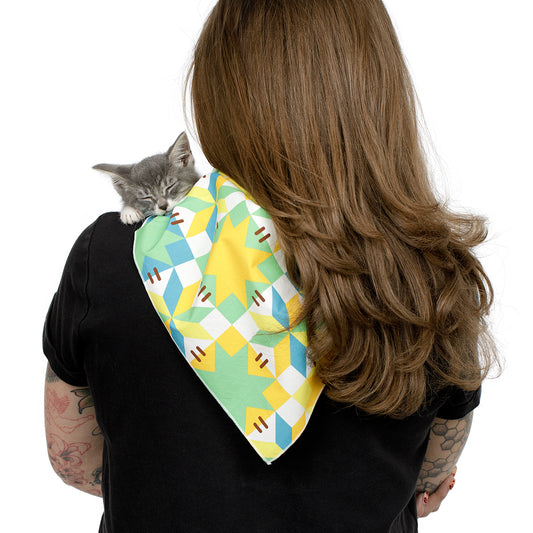 A woman with long brown hair seen from behind, wearing a black shirt and carrying a small gray kitten on her shoulder. The kitten is wrapped in a colorful patterned bandana with yellow, green, blue, and white geometric shapes.