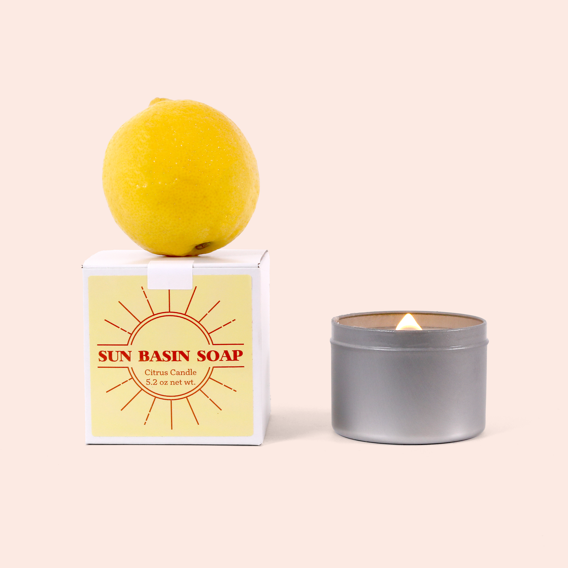 The same lit candle in its gray tin next to its white product box labeled "SUN BASIN SOAP Citrus Candle 5.2 oz net wt." with a red sun logo design, topped with a yellow lemon, all arranged against a pink background.