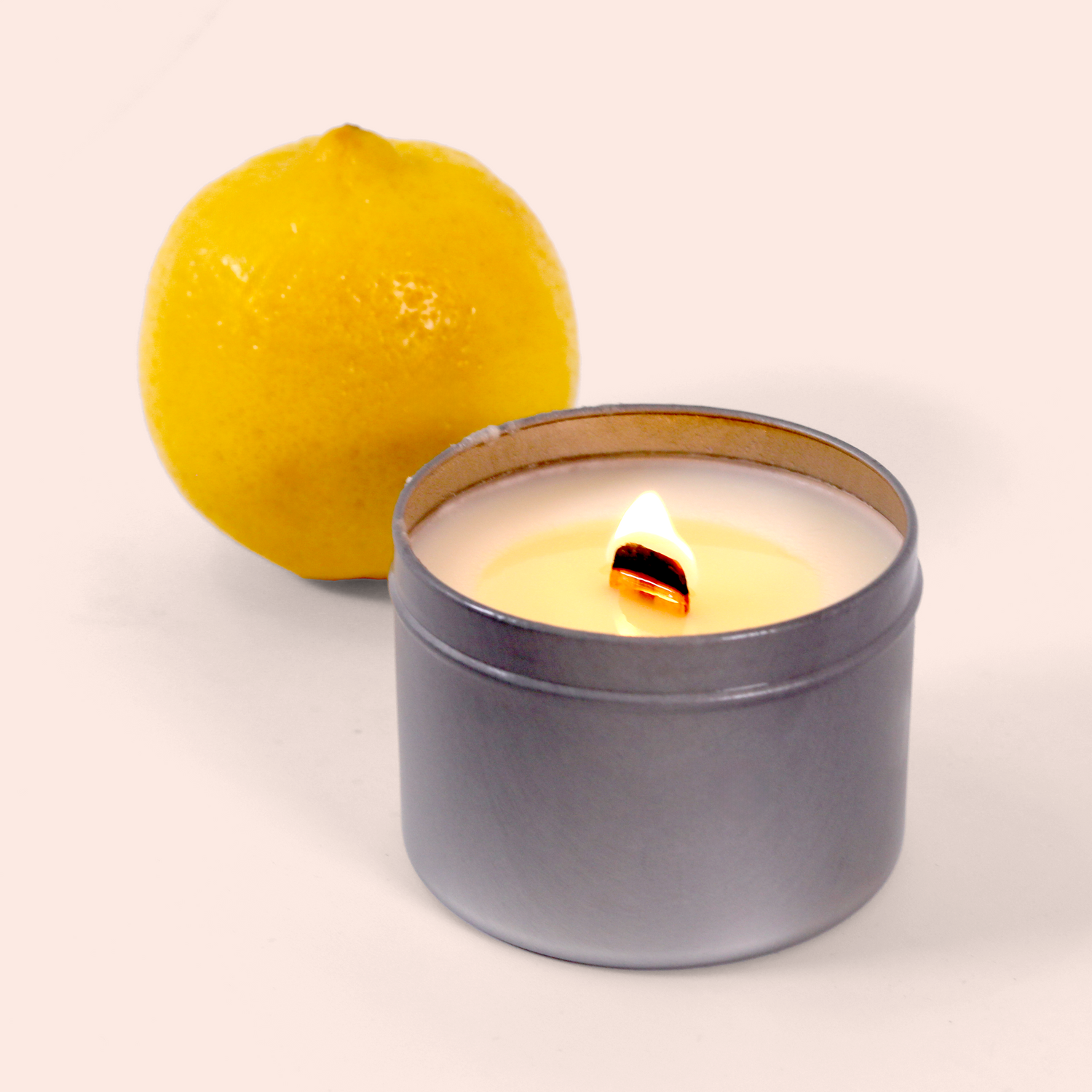  A lit candle in a gray metal tin container with cream-colored wax and a wooden wick, positioned next to a fresh yellow lemon against a soft pink background.