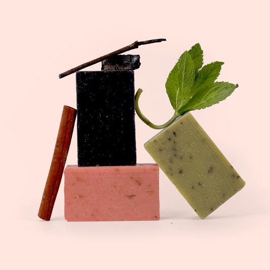 An artistic arrangement of handmade soaps in different colors with foamy bubbles. The image shows three bars of soap - a black rectangular bar on top of a pink bar, with a green bar leaning against them. The black and green soaps have foamy bubbles on them, and the green soap has fresh mint leaves attached. There are also cinnamon sticks positioned alongside the soaps. The composition is set against a light pink background.