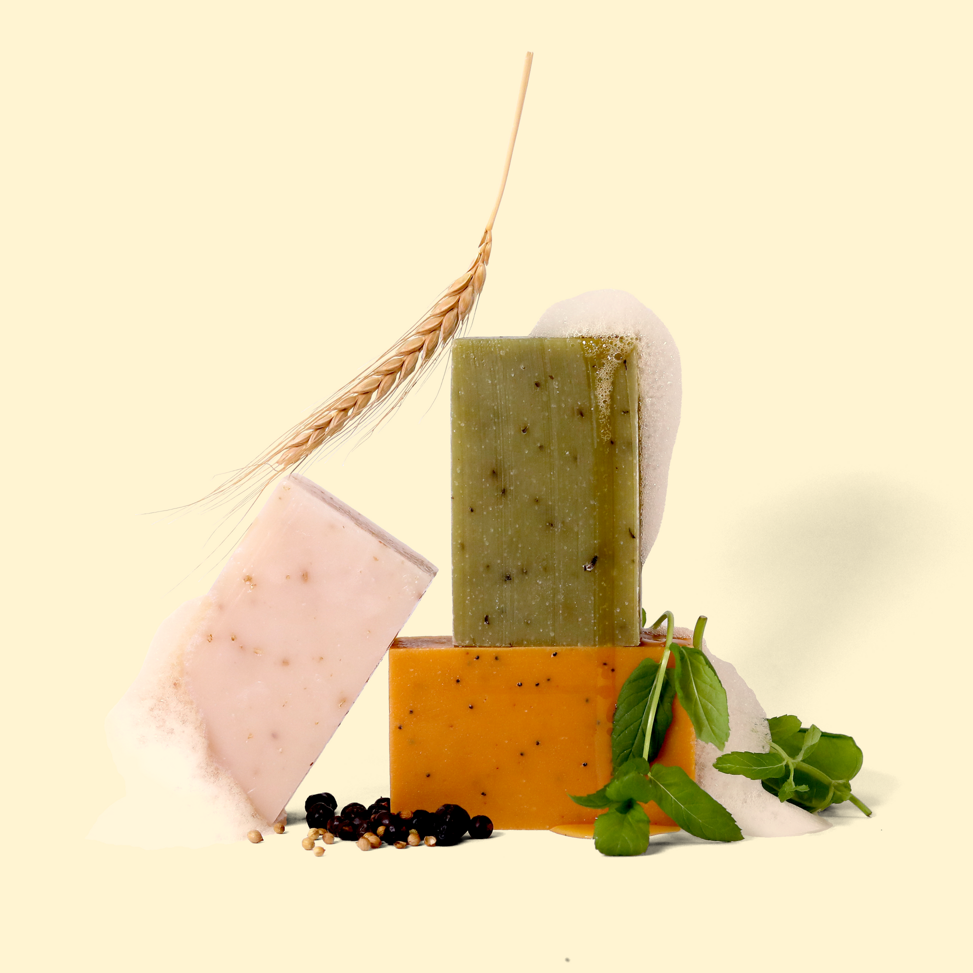 Arrangement of three soap bars in different colors (green, orange, pink) with natural elements. A wheat stalk leans against the soaps, while herbs and coffee beans are scattered at the base. The soaps are on a light cream background. Sun Basin Soap - Good Store.