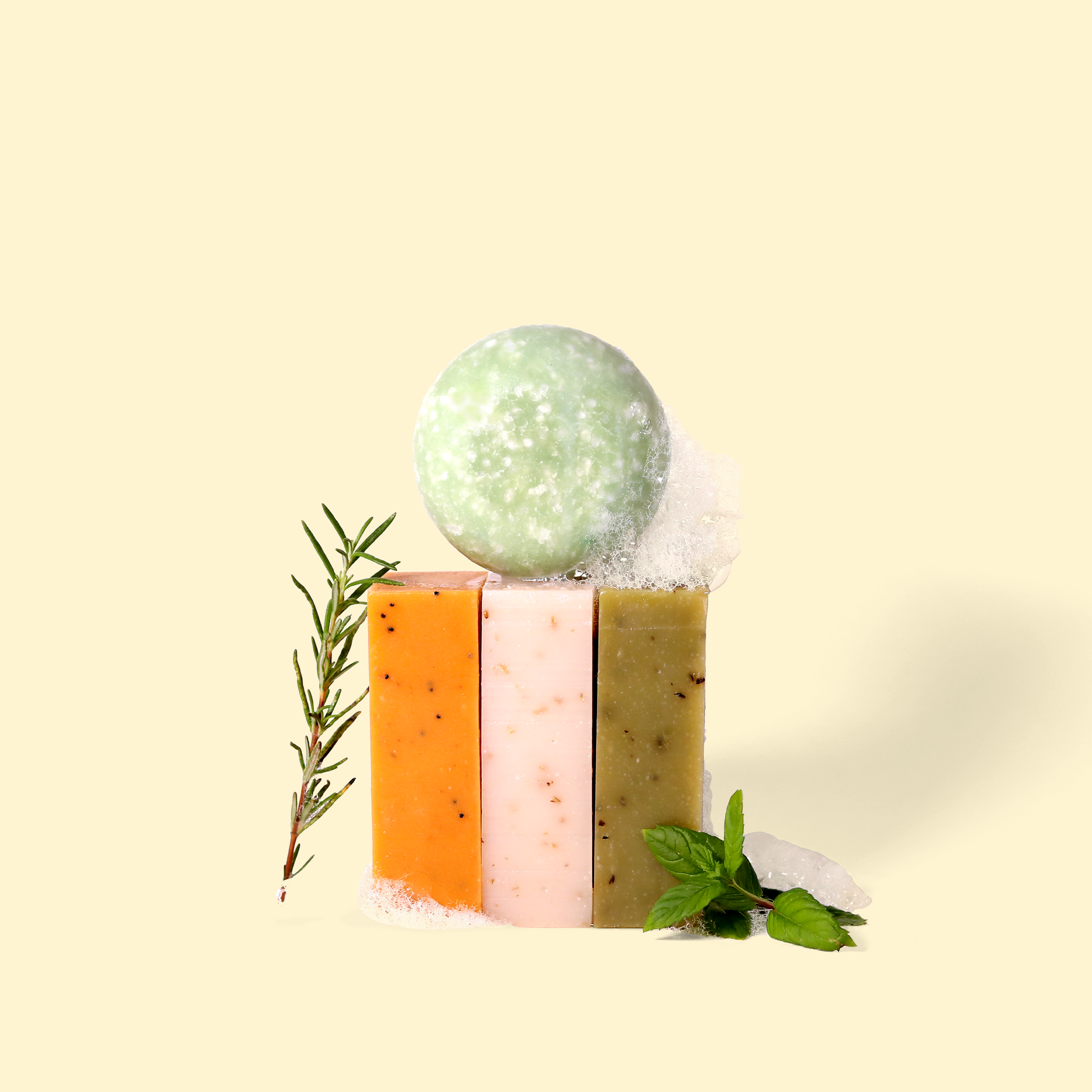 A round pale green shampoo bar sits atop three rectangular bars in orange, pink, and olive green. A sprig of rosemary leans against the left side, while fresh mint leaves rest on the right. White foam accents the composition and it is set against a cream background. Sun Basin Soap - Good Store. 