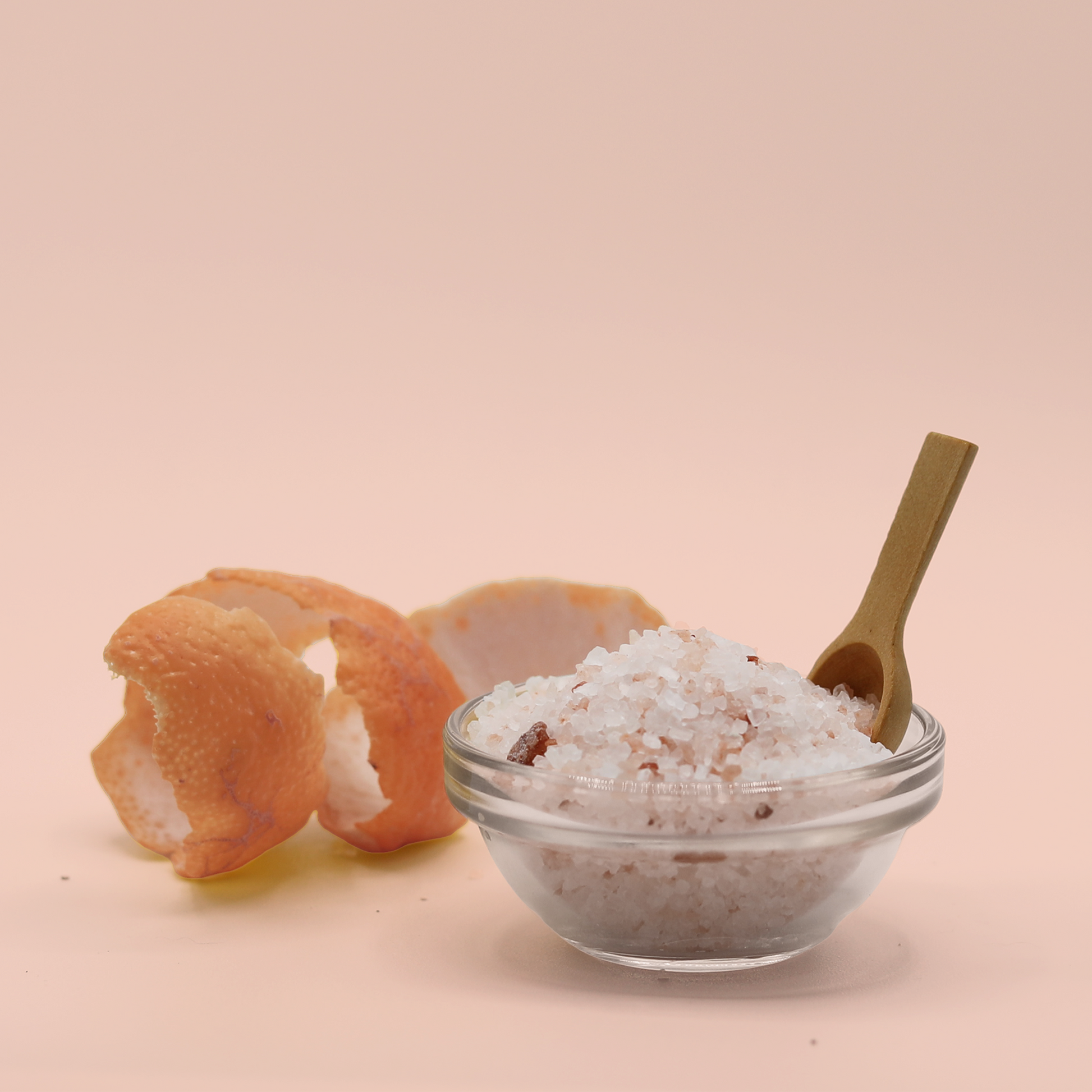 A little glass bowl filled with light pink bath salts next to a curled grapefruit peel. A Good Store product.