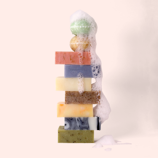 A stack of colorful soap bars with foam cascading down one side. From top to bottom: a green spherical shampoo bar, a yellow spherical shampoo bar, pink, purple, cream, brown speckled, orange-yellow marbled, black and white marbled, and green herb-speckled rectangular soap bars. Sun Basin Soap - Good Store.