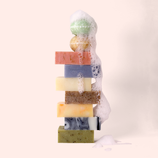 A stack of colorful soap bars with foam cascading down one side. From top to bottom: a green spherical shampoo bar, a yellow spherical shampoo bar, pink, purple, cream, brown speckled, orange-yellow marbled, black and white marbled, and green herb-speckled rectangular soap bars. Sun Basin Soap - Good Store. 