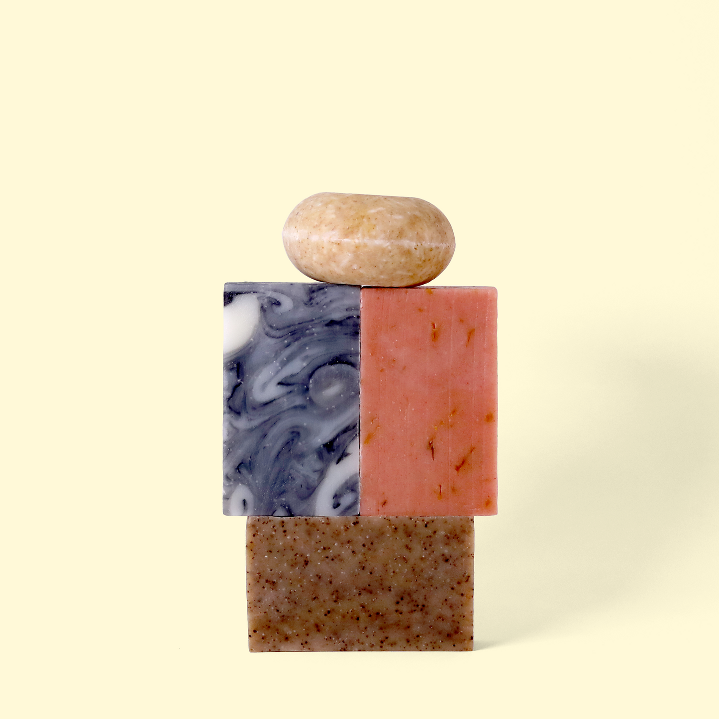 Four artisanal soap bars stacked in a tower against a cream background. From bottom to top: a speckled brown bar, a marbled blue and white bar next to a pink bar, and a rounded beige bar on top.