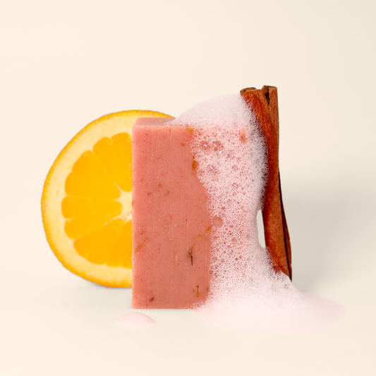 A rectangular block of pink Daydreamer soap standing upright, with a slice of orange to its left and a cinnamon stick to its right. The soap is partially covered in white soap lather, spreading from right to left across the soap's surface. The soap is set against a light background. Sun Basin Soap - Good Store. 