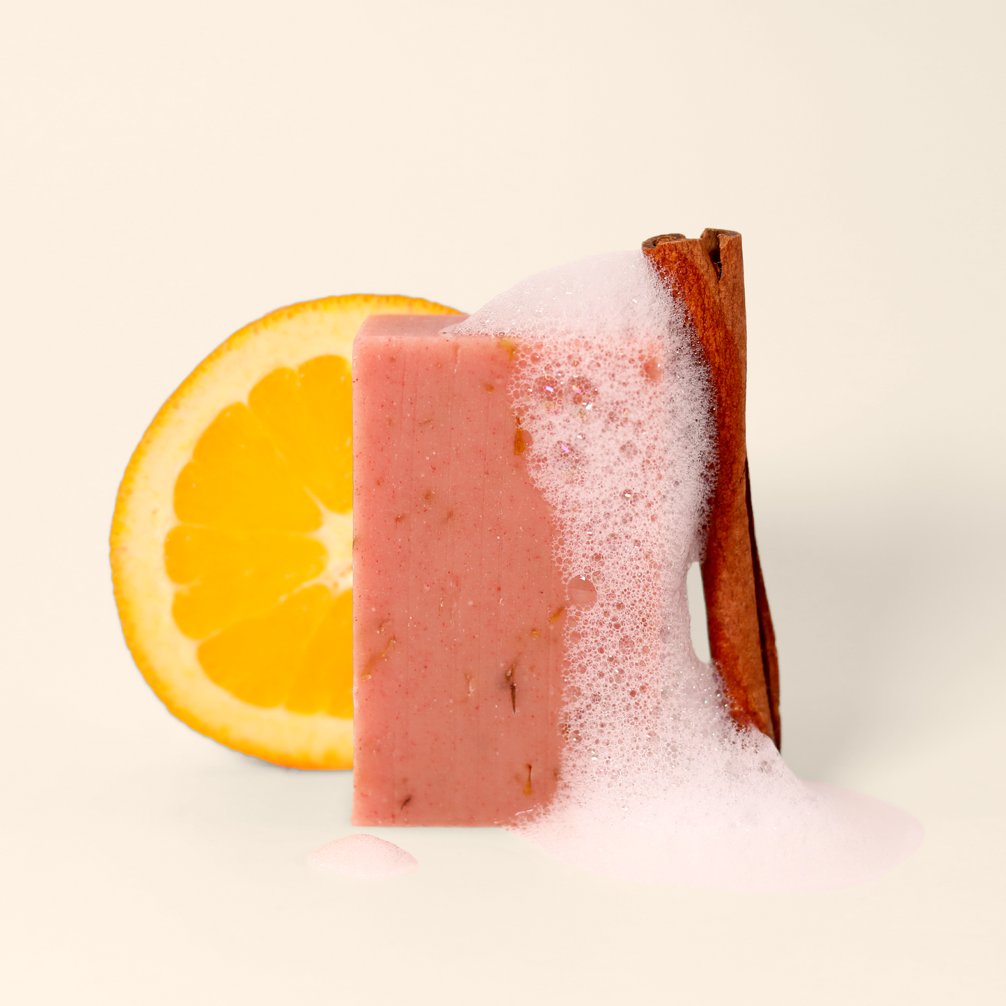 A close-up of a pink bar of soap with foamy lather and a cinnamon stick leaning against it. Behind the soap is a slice of orange. The soap appears to have small specks or botanical elements embedded within it. The image is set against a neutral light background, highlighting the contrast between the pink soap and the white foam.