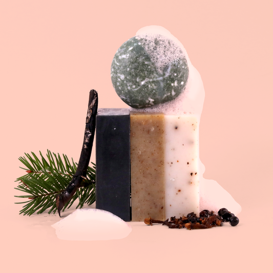 An artful arrangement of bath products including a green bath bomb, black charcoal soap bar, beige speckled soap bar, and white soap bar, styled with a pine branch, dried spices, and white foam elements against a peachy pink background. The products are stacked to create a sculptural composition.