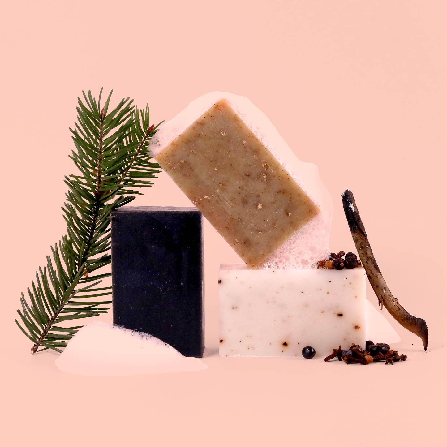 An artistic arrangement of three soap bars (white, beige, and black) with a pine branch, dried spices, and a wooden stick against a pink background. 