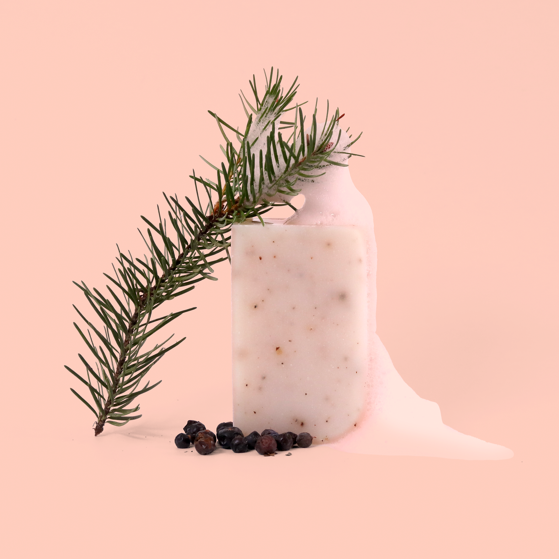 A natural bar of white soap with visible speckles displayed against a pale pink background, accompanied by a pine branch and scattered juniper berries. The composition is minimalist and styled like a product photo.